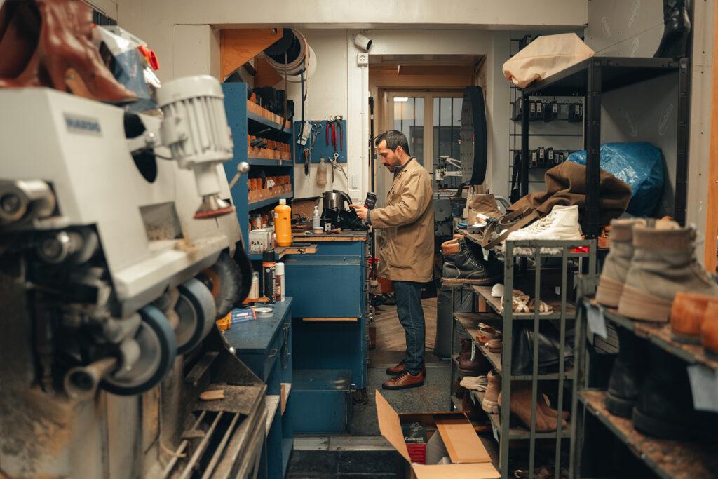 Artisan cordonnier dans son atelier à Neuchâtel, entouré d’étagères remplies de chaussures et de matériel de réparation.
