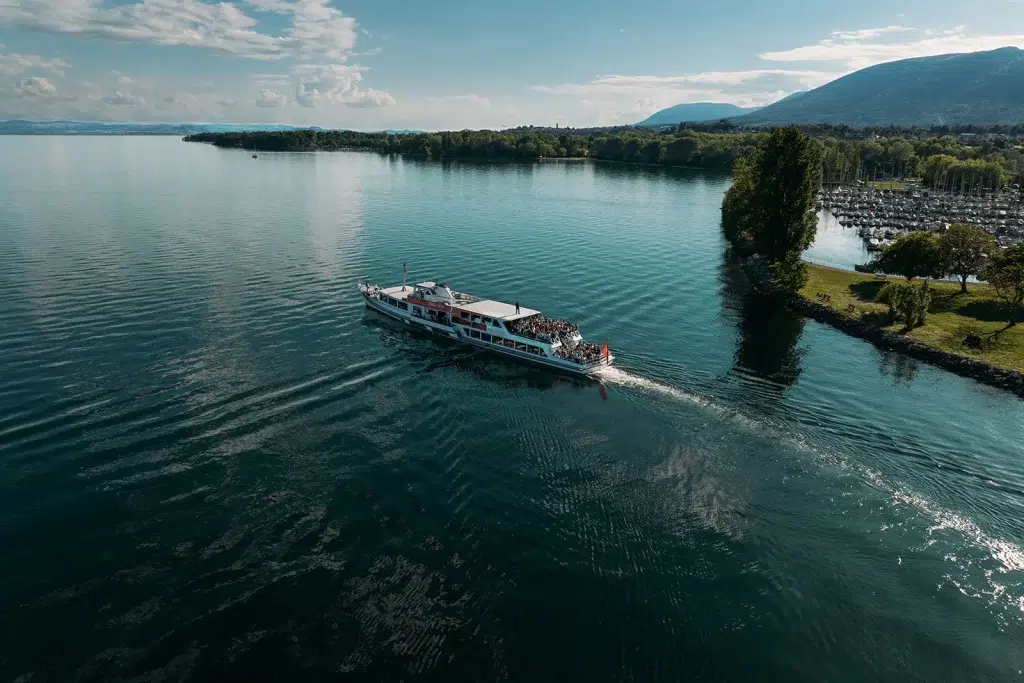 Bateau de croisière de LNM et Vin & Terroir, naviguant sur le lac de Neuchâtel entouré de montagnes et d’arbres par une journée ensoleillée pendant les caves ouvertes 2025, capturé en vue aérienne.